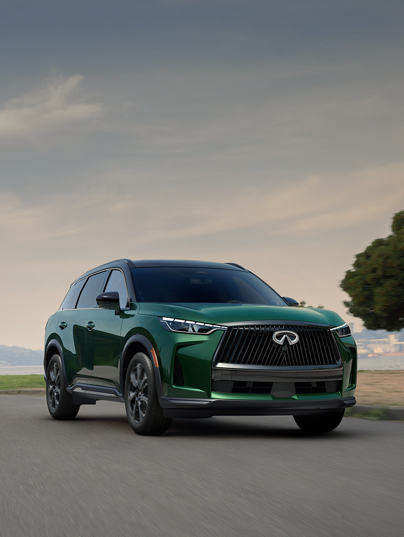 Front view of a green Infiniti SUV driving along a scenic road, with trees and water in the background.