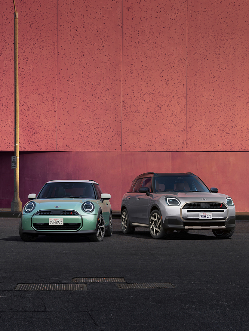 Two MINI vehicles—a mint-green MINI Cooper and a silver MINI Countryman—parked side by side in front of a modern pink building.