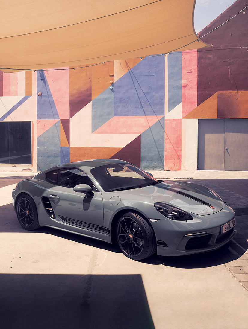 Porsche 718 Cayman parked in a sunlit urban courtyard with geometric mural walls in the background.