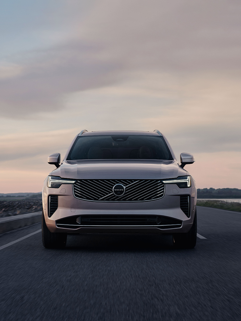 Front view of a gray Volvo SUV driving on a coastal road at sunset, with a serene water body on the side.